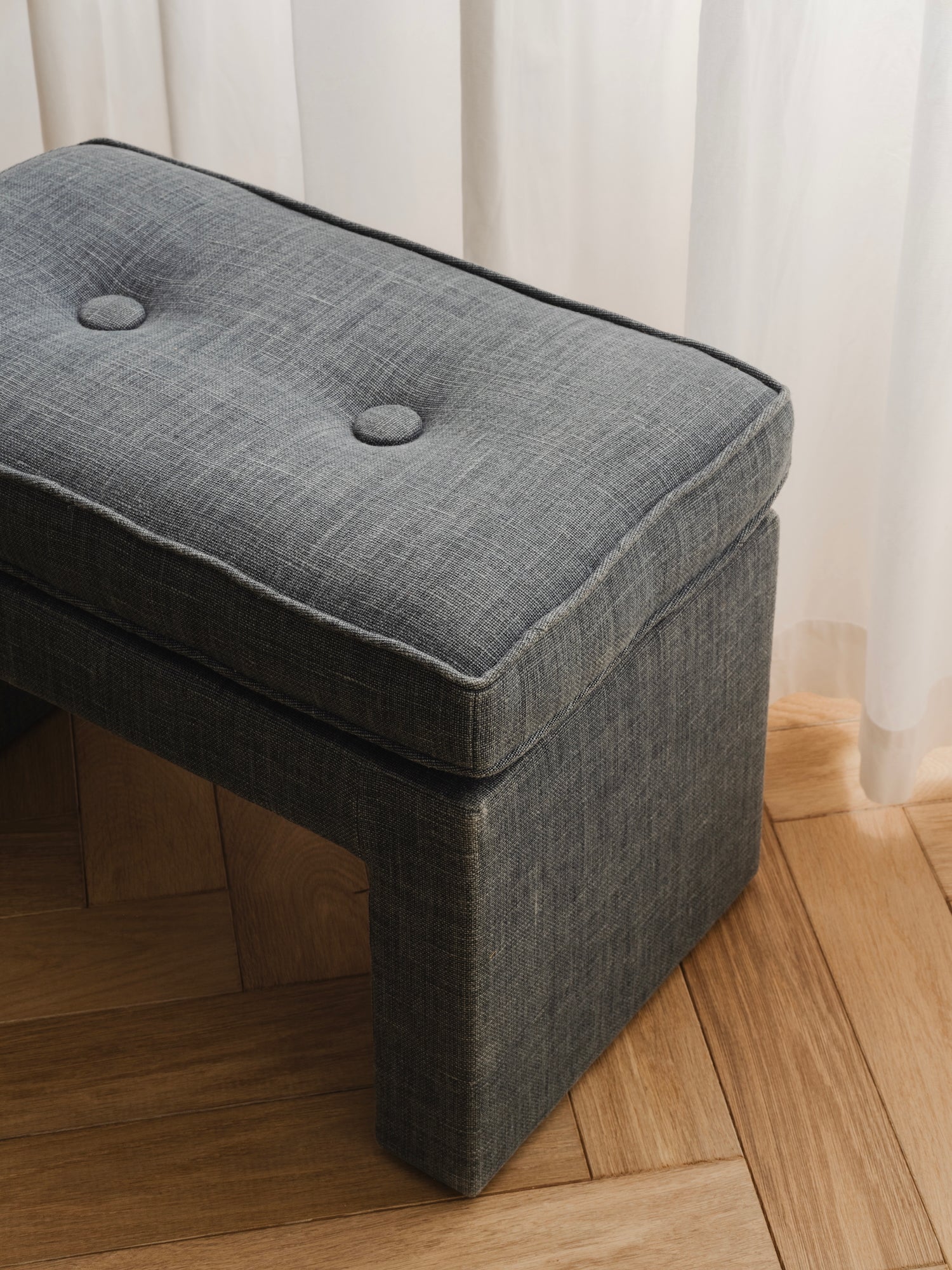 Blue upholstered ottoman on a wooden floor with white curtains in the background