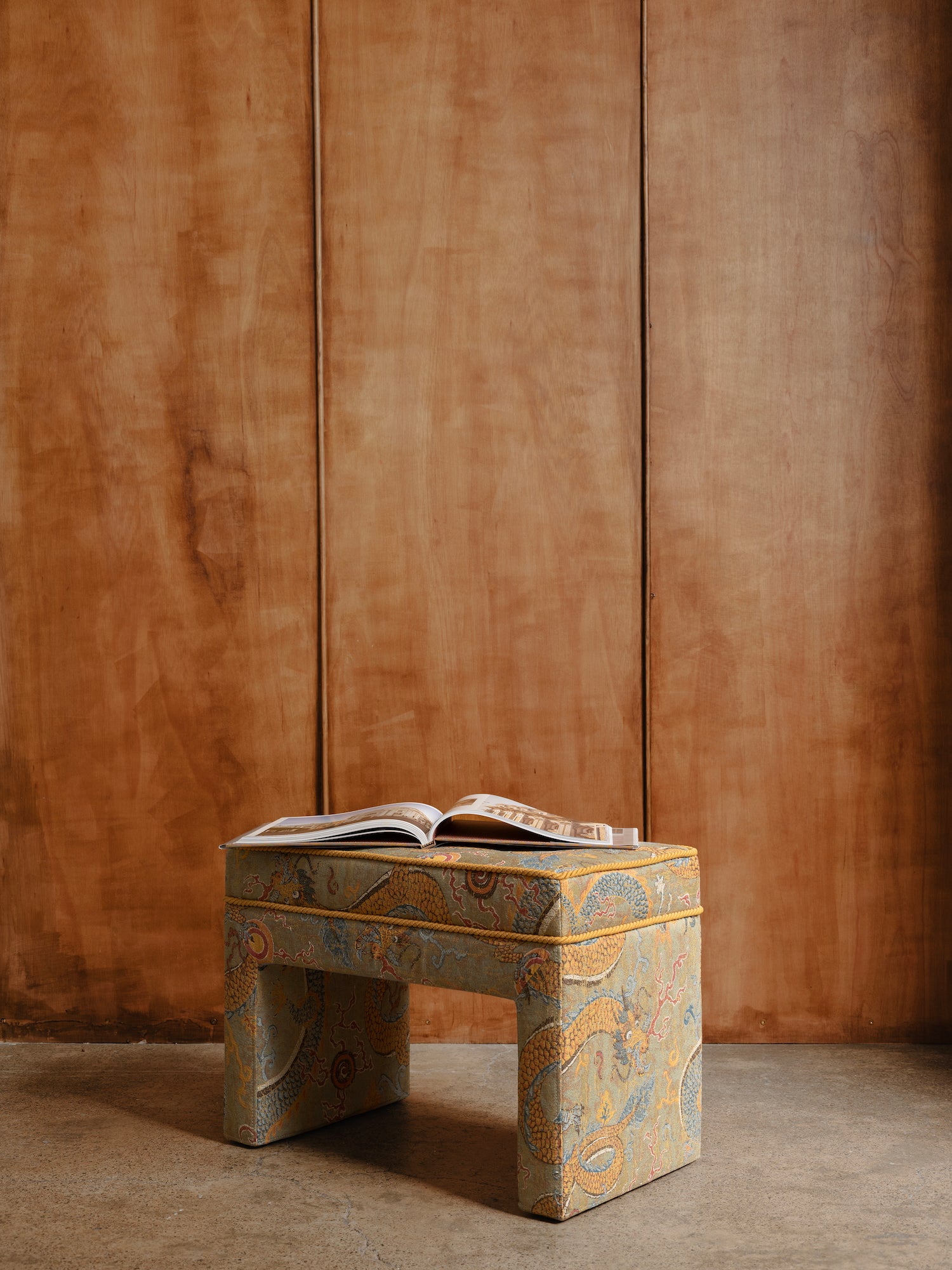 Upholstered stool with an open book on a cement floor against a wood panelled wall.