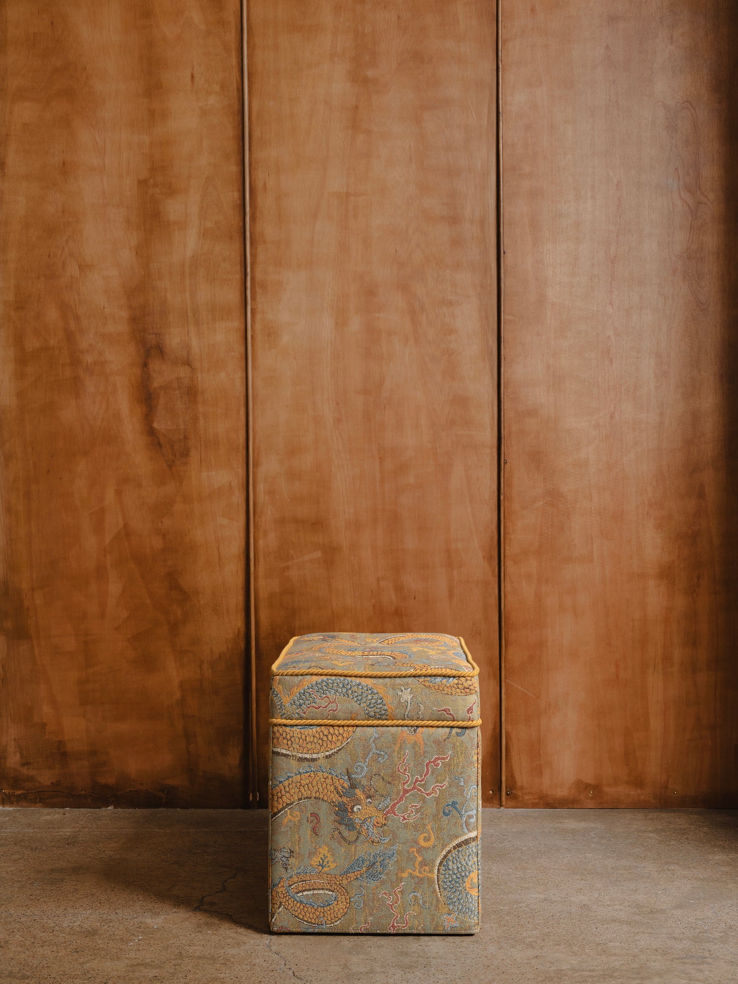 Upholstered stool with intricate patterned fabric against a wood panelled wall