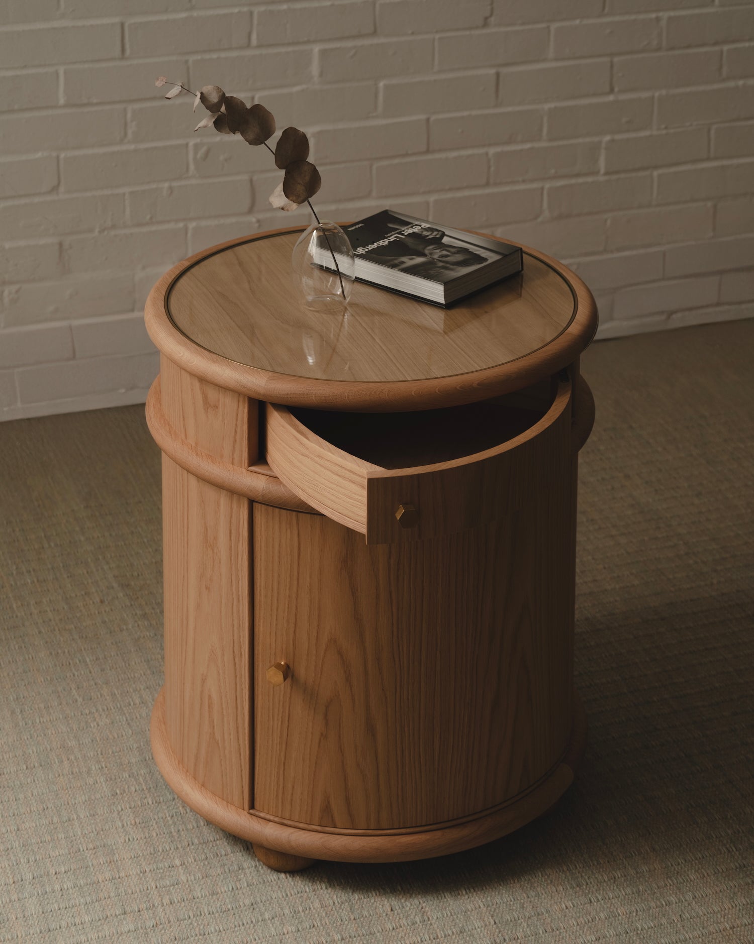 Wooden bedside table with glass top, brass handles and a drawer against a brick wall.